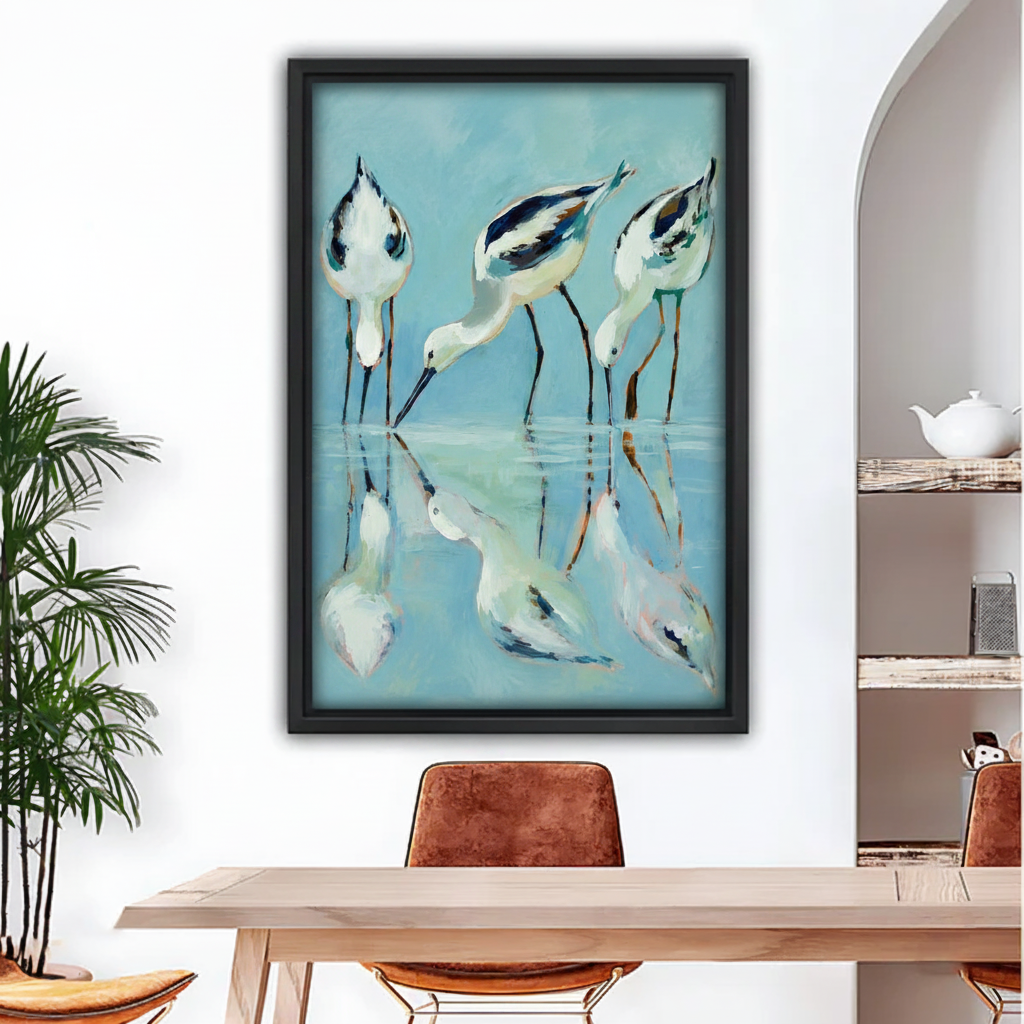 Three avocet shorebirds wading water reflection framed wall art print, black frame, teal blue coastal oil painting style, hung above a light wood dining table with leather chairs in a bright modern dining room with indoor palm