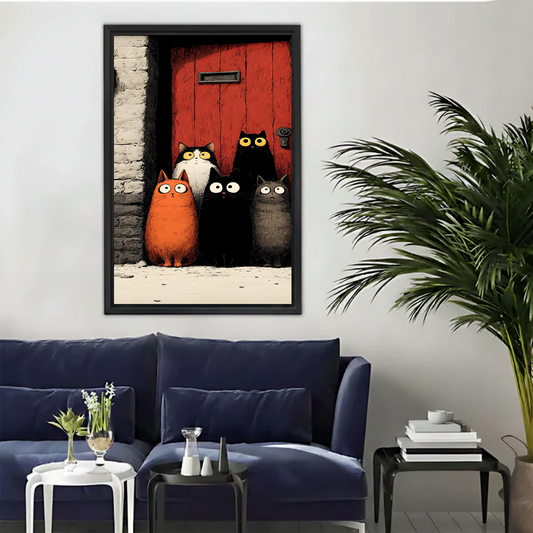 Five wide-eyed cartoon cats orange black-white small black large black and grey crowded at red wooden door gothic illustration framed wall art hung on white wall above navy blue sofa with white side tables and palm plant in modern living room