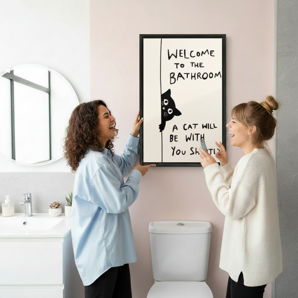 Two women hanging funny cat quote framed print in pink bathroom - humorous black cat bathroom canvas wall decor