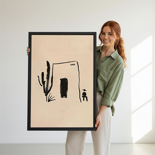 Woman holding black-framed desert house and cactus canvas wall art featuring a dark doorway and small seated figure.