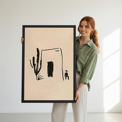 Woman holding black-framed desert house and cactus canvas wall art featuring a dark doorway and small seated figure.