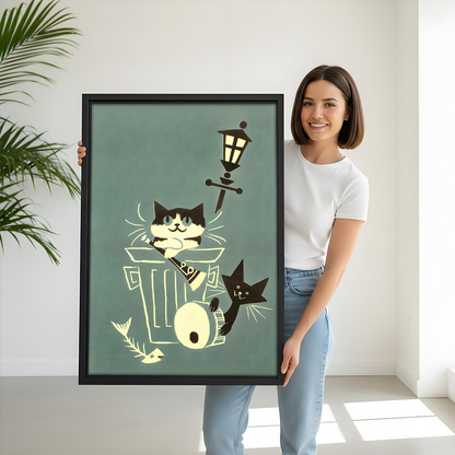 Woman holding black-framed alley cats canvas wall art showing two playful cats around a trash can beneath a streetlamp.