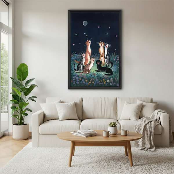 Framed whimsical moonlit cats canvas wall art with a navy sky, displayed beside green houseplants and wooden tables.