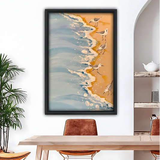 Sandpiper shorebirds on sandy beach with ocean waves framed wall art print, black frame, warm golden sand coastal oil painting style, hung above a light wood dining table with leather chairs in a bright modern dining room with indoor palm