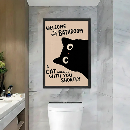 Black-framed black cat bathroom canvas wall art with a funny welcome quote above a toilet against marble walls.