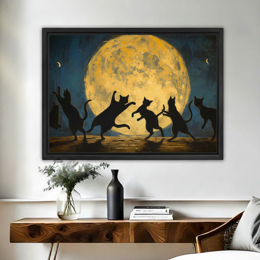 Black framed dramatic black cat moonlight dance canvas print hung above a rustic walnut console table in a bright living room with a white floral arrangement, black ceramic vase, and stacked books beside a linen armchair