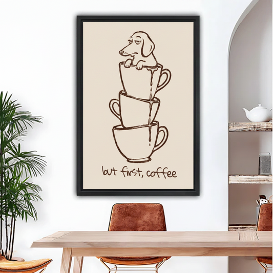 Black framed wall print featuring a cartoon dachshund perched in stacked mugs with But First Coffee text, hung in a modern beige dining space