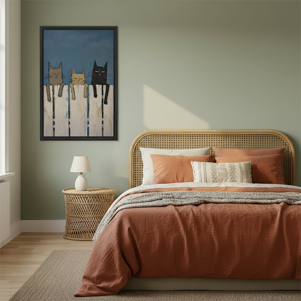 Framed folk art style wall print of three cats on white fence displayed in cozy bedroom with sage green walls and terracotta bedding