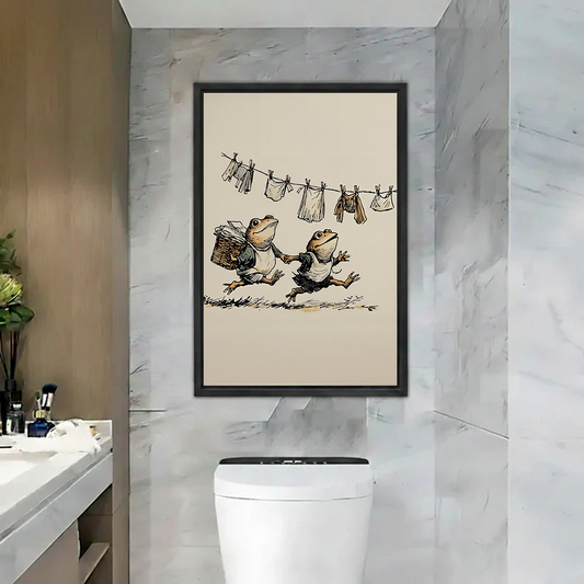 Black-framed frog laundry canvas wall art showing two frogs beneath a clothesline displayed above a bathroom toilet.