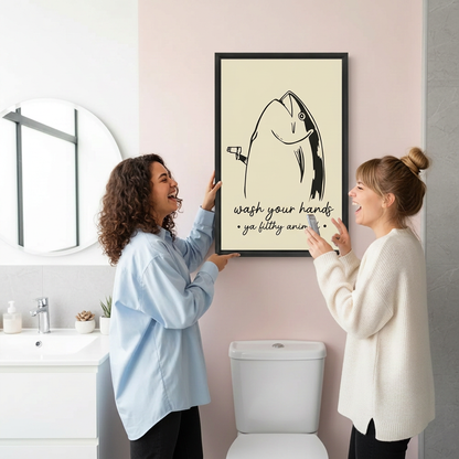 Two women laughing beside black-framed funny fish wall art displayed on a pink bathroom wall.