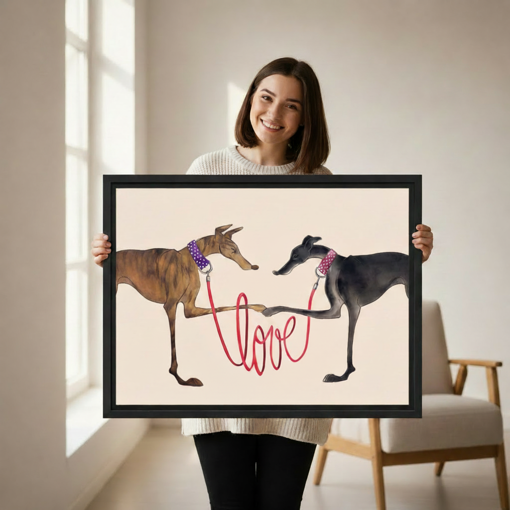Woman holding large framed two greyhounds with red love leash wall art print, black frame, whimsical dog love illustration cream background, showing actual print size in a bright modern studio