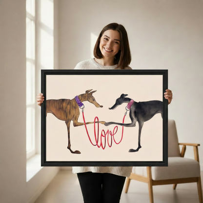 Woman holding large framed two greyhounds with red love leash wall art print, black frame, whimsical dog love illustration cream background, showing actual print size in a bright modern studio