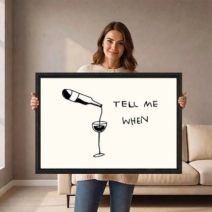 Tell Me When Wine artwork held in a black frame by a woman beside a cream sofa.