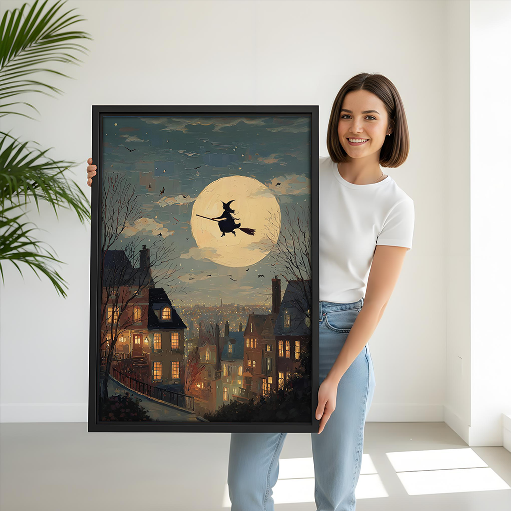 Woman holding black-framed moonlit witch canvas wall art showing a flying witch above a village with glowing windows.