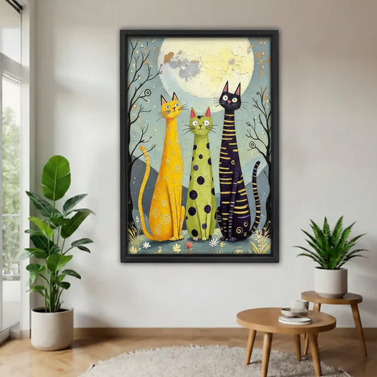 Large black framed print of three tall quirky cats in bold patterns standing in a moonlit meadow with wildflowers, hung on a white wall flanked by tropical potted plants