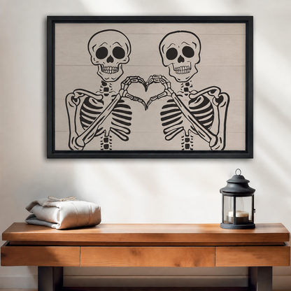 Two skeletons making a heart with their hands on shiplap background framed canvas above warm wood console table with black lantern and folded linen