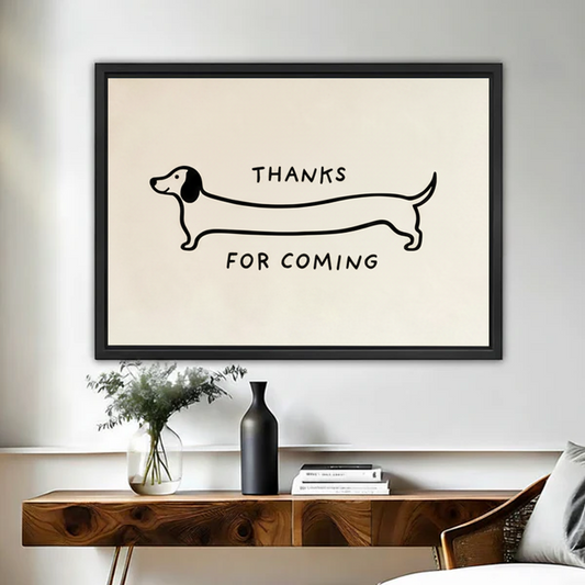 Black framed minimalist dachshund line art canvas with the humorous quote Thanks For Coming, featuring a single continuous black line drawing of an extra-long sausage dog on a cream background, displayed above a walnut console table with a black vase and greenery