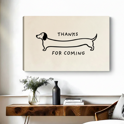 Frameless gallery-wrap canvas of a minimalist one-line dachshund illustration with Thanks For Coming text in a hand-lettered style on a warm cream background, hung above a walnut console table with a black ceramic vase and stacked books