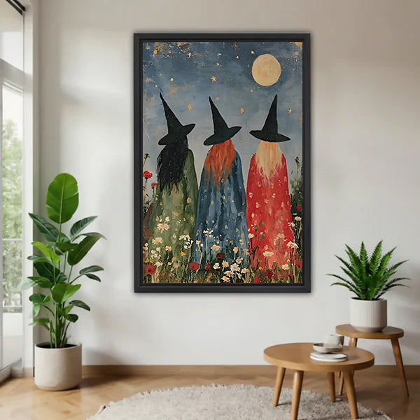 Three witches moonlit wildflower folk art green blue red robes black hats gold star full moon daisy poppy black framed canvas wall art on white wall beside round wood side table with monstera and fern plant