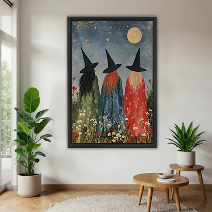 Three witches moonlit wildflower folk art green blue red robes black hats gold star full moon daisy poppy black framed canvas wall art on white wall beside round wood side table with monstera and fern plant