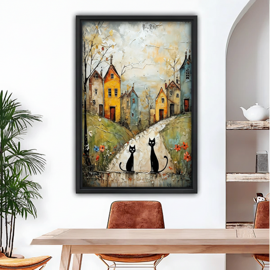 Black framed oil-style wall art of two sleek black cats on a cobblestone village road flanked by yellow and blue townhouses with red wildflowers, hung above a rustic dining table in a bright arched interior