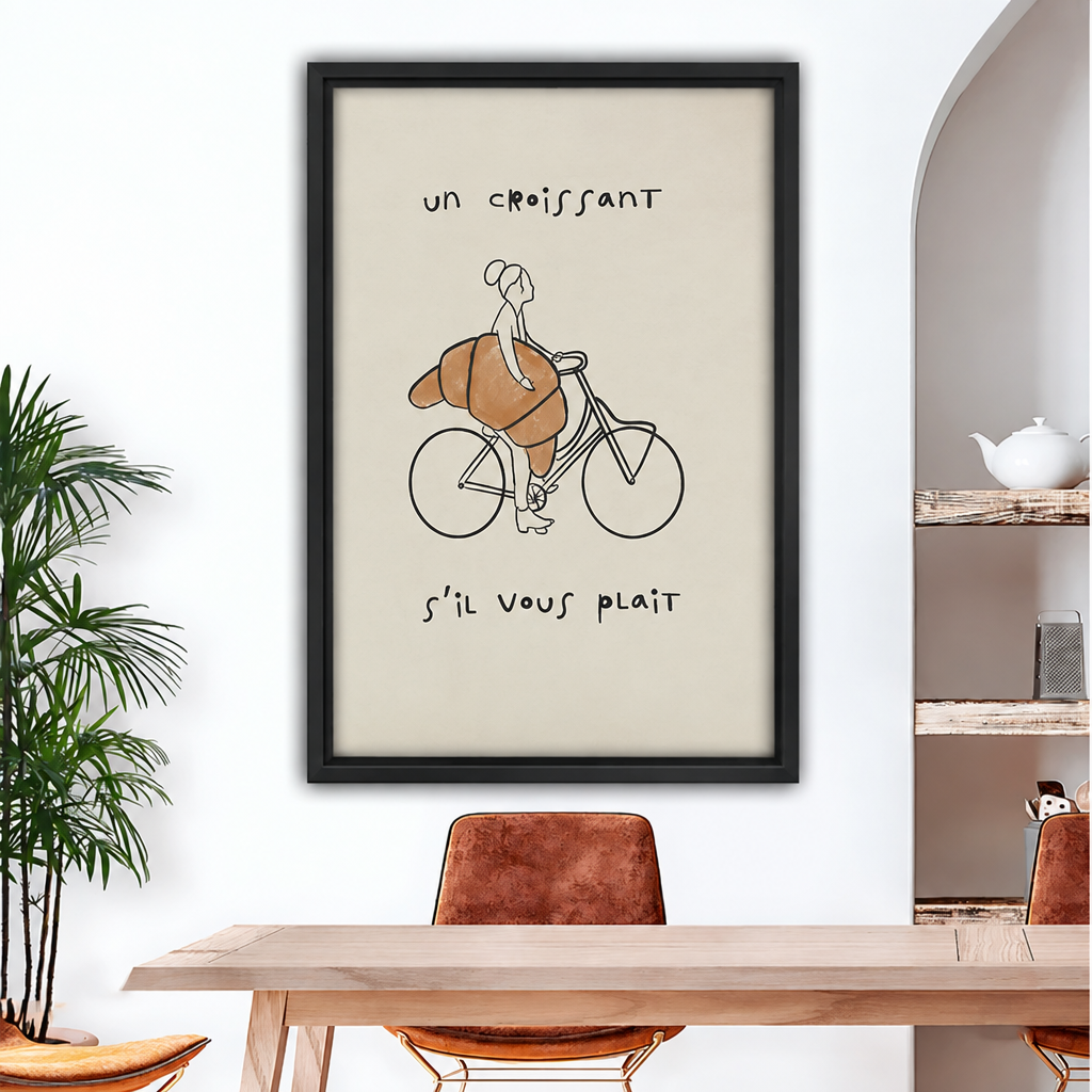 Framed canvas of minimalist line art of a woman riding a bicycle with a giant golden croissant and French text "un croissant s'il vous plait" on cream background above wood dining table with brown leather chairs