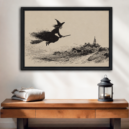 Black-framed vintage flying witch wall art displayed above a wooden console beside folded linens and a candle lantern.