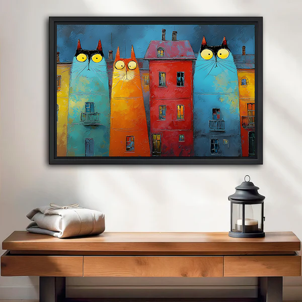 Black-framed whimsical cat buildings canvas wall art in turquoise, orange, and red above a wooden console beside a lantern.