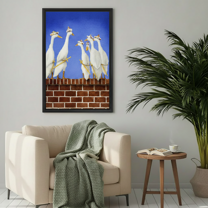 Humorous framed canvas print of white geese in party hats perched on a red brick wall under a bright blue sky, hung above a cream armchair with a green knit throw