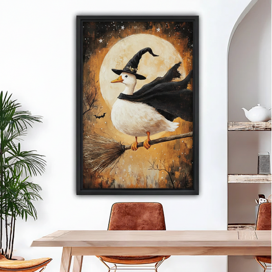 Black framed oil-style Halloween wall art of a cute white goose wearing a witch hat and flowing black cape on a broomstick with bats and stars, hung above a rustic dining table